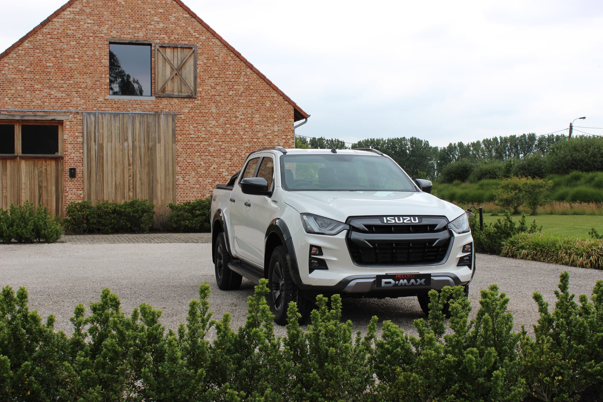 White ISUZU D-MAX parked on a gravel driveway, in front of a rustic brick and wooden building, surrounded by greenery.
