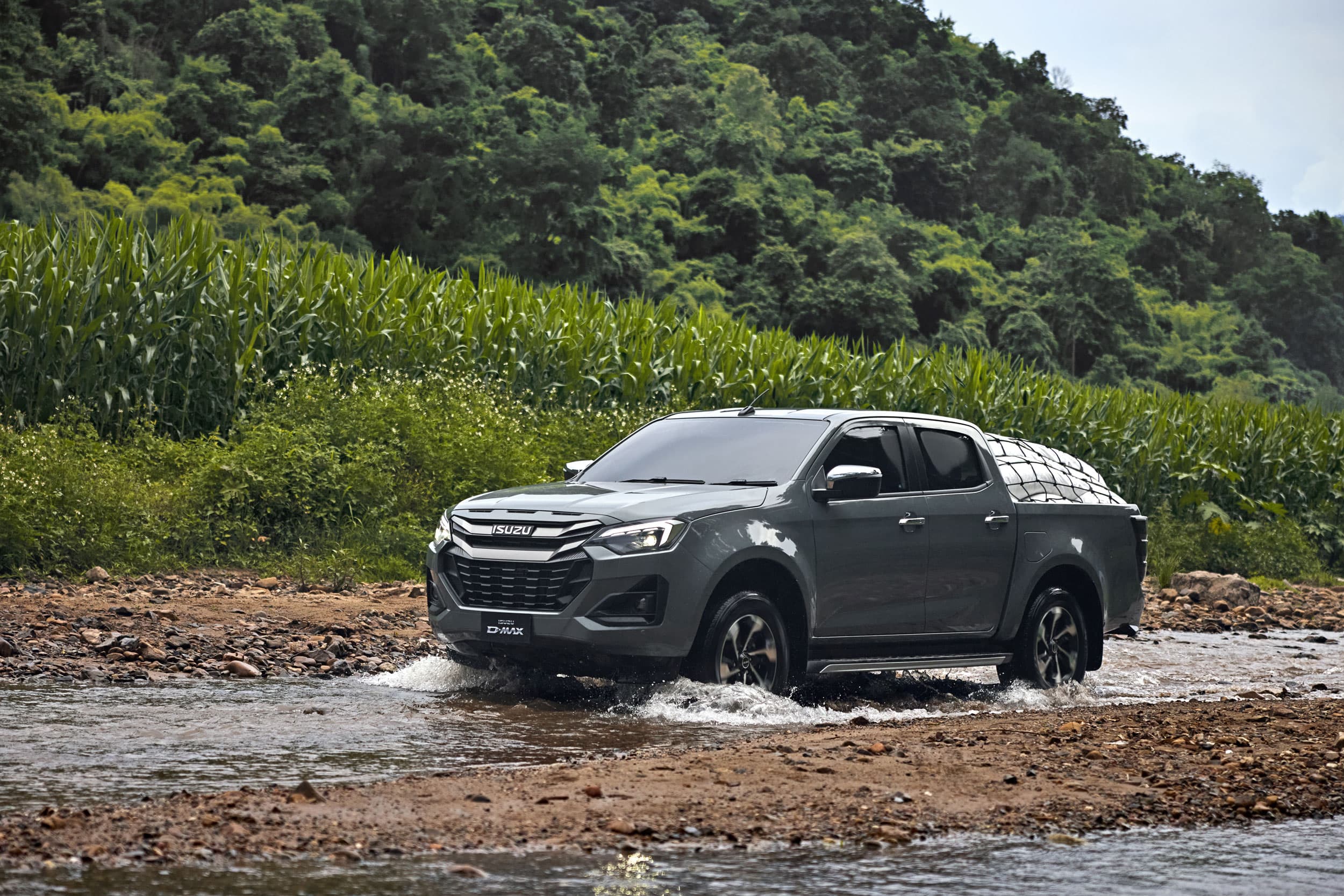A gray ISUZU D-MAX drives through a shallow stream surrounded by lush green vegetation and trees.
