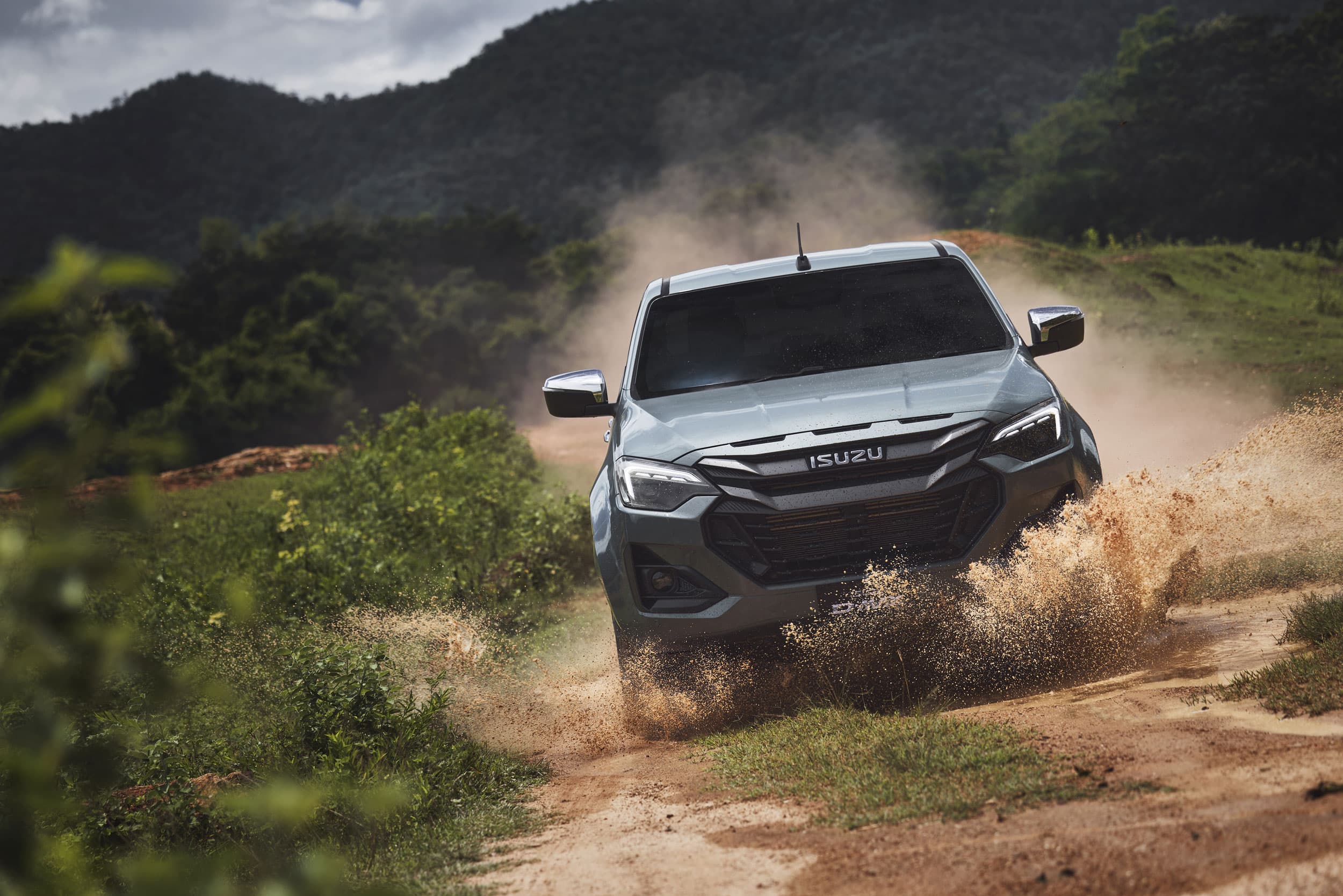 A silver ISUZU D-MAX truck drives through a muddy off-road trail, kicking up dust and water, set against a backdrop of lush greenery and hills.