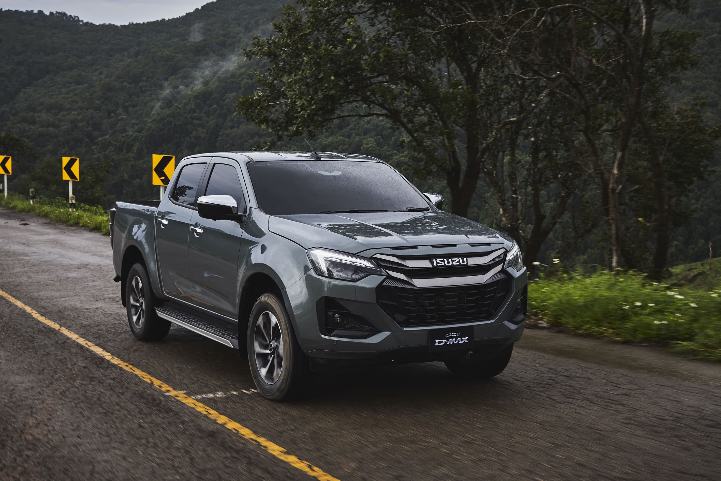 A gray ISUZU D-MAX pickup truck driving on a wet road with lush green trees and hills in the background.