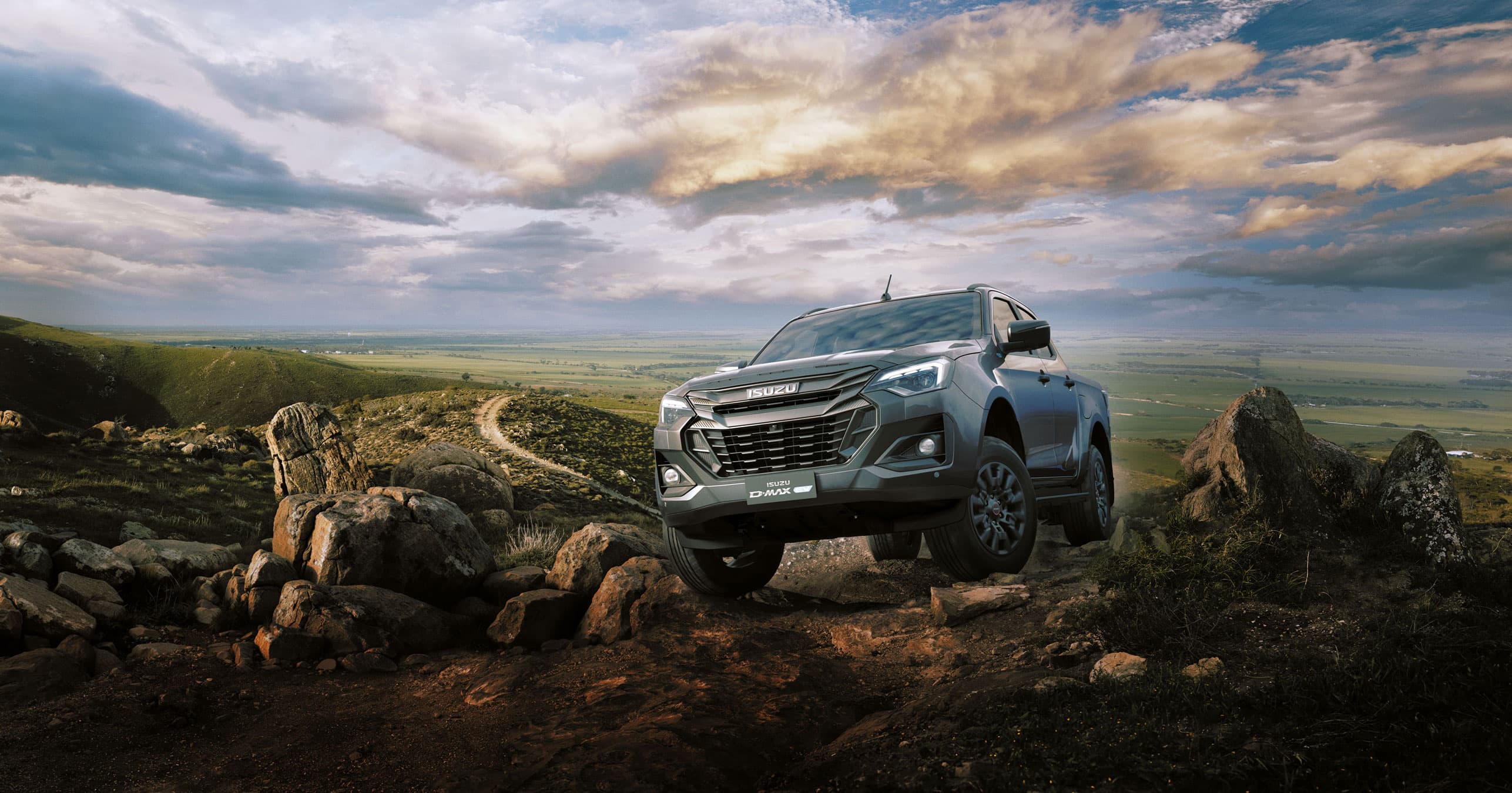 ISUZU D-MAX EV driving on a rocky terrain against a vast, cloudy landscape under a dramatic sky.