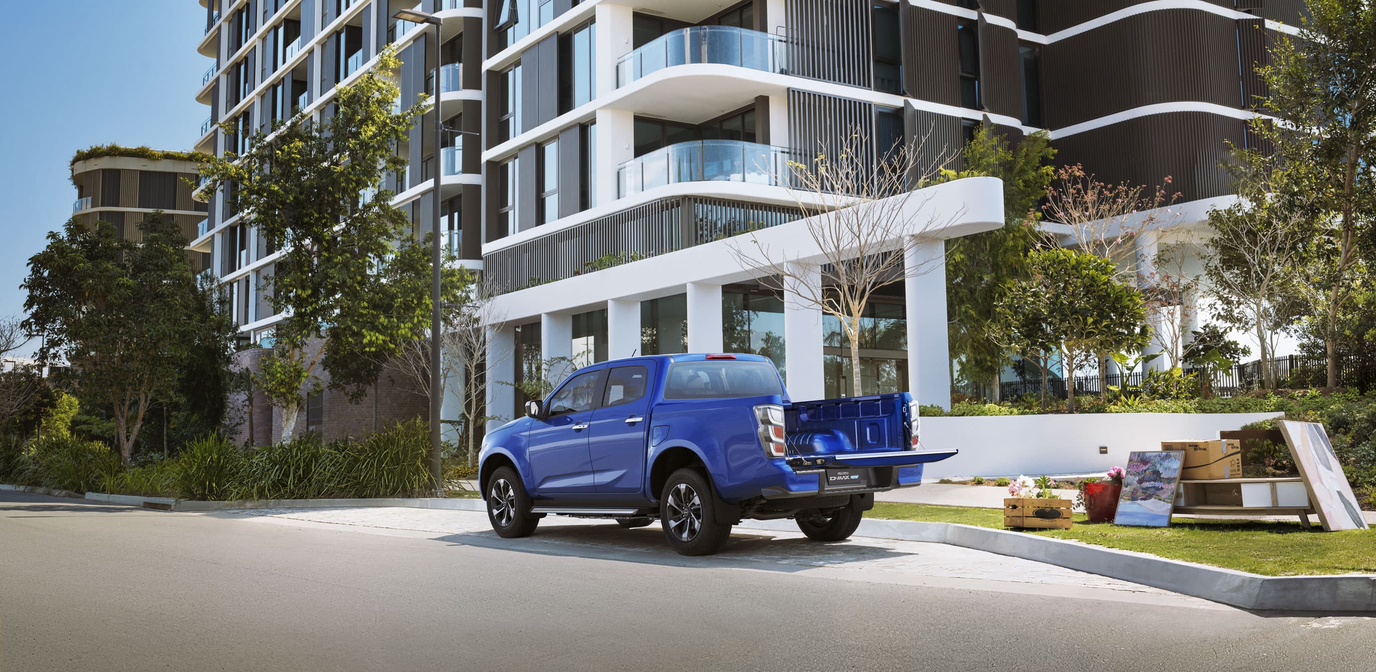 Blue ISUZU D-MAX EV parked outside a modern apartment building, with various items on the pavement nearby.
