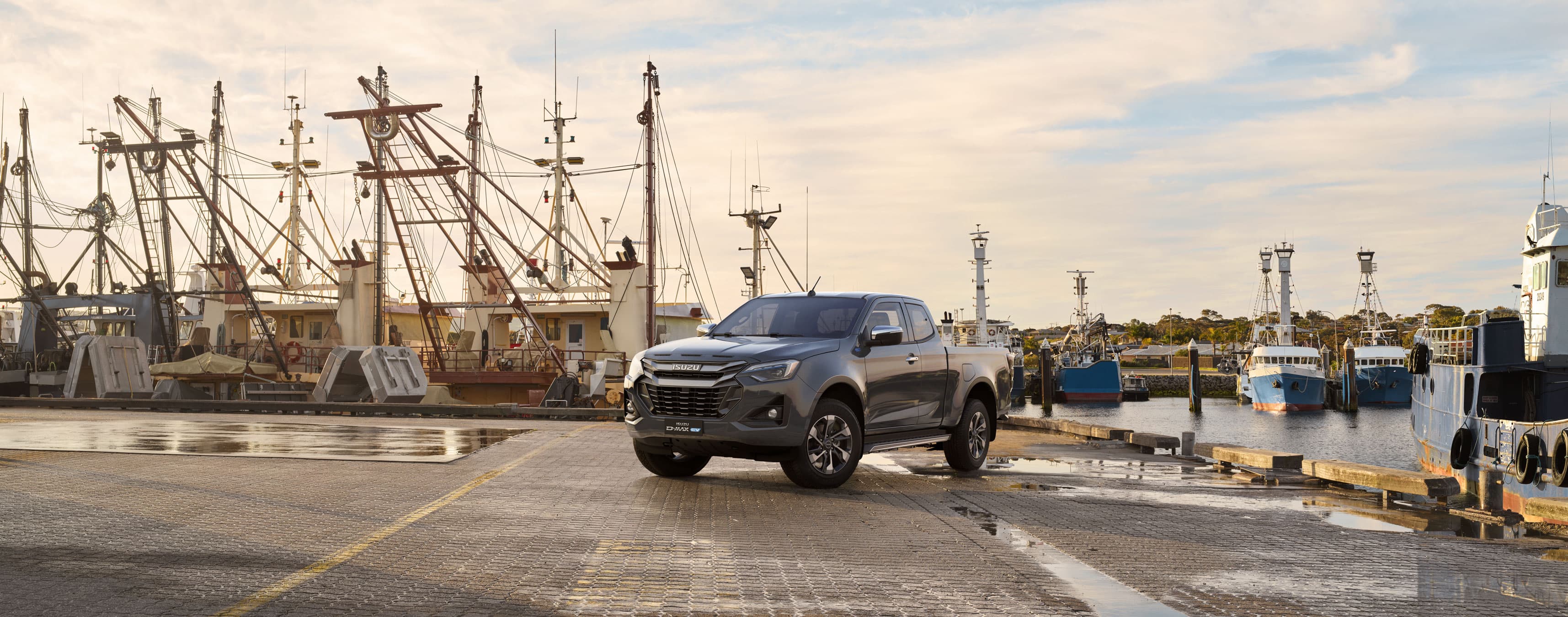 A dark gray ISUZU D-MAX EV is parked on a dock, with several fishing boats moored in the background under a cloudy blue sky.