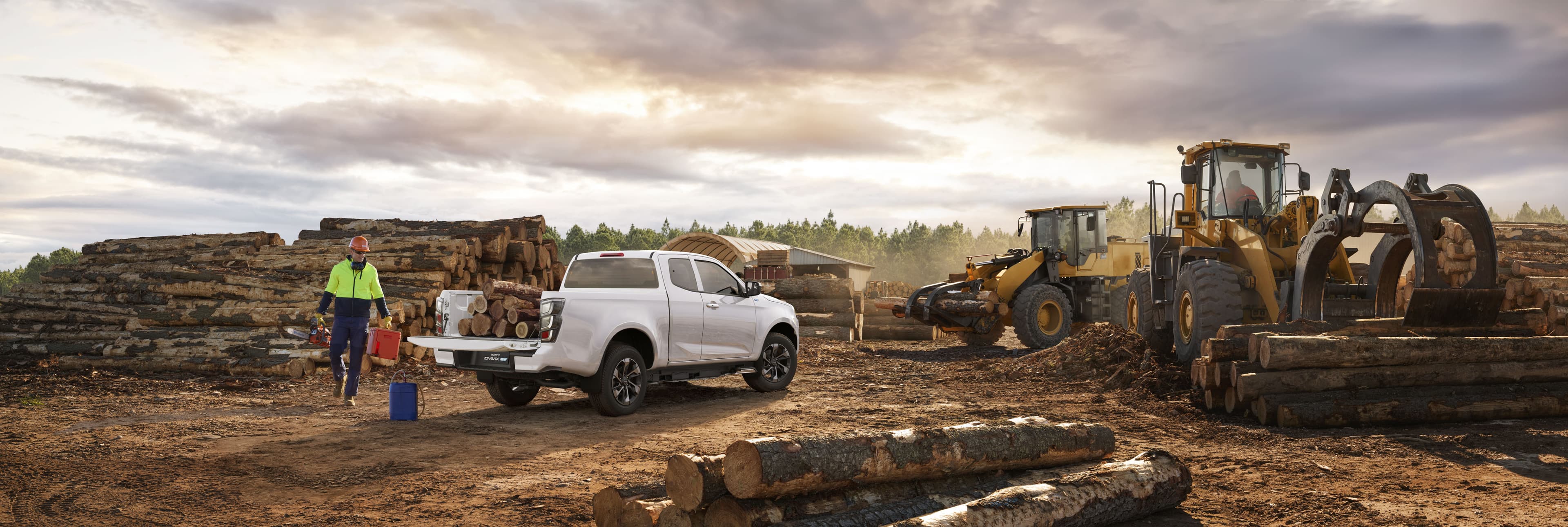 A white ISUZU D-MAX EV loaded with firewood is parked at a lumber yard. Two workers and heavy machinery are seen handling large logs.
